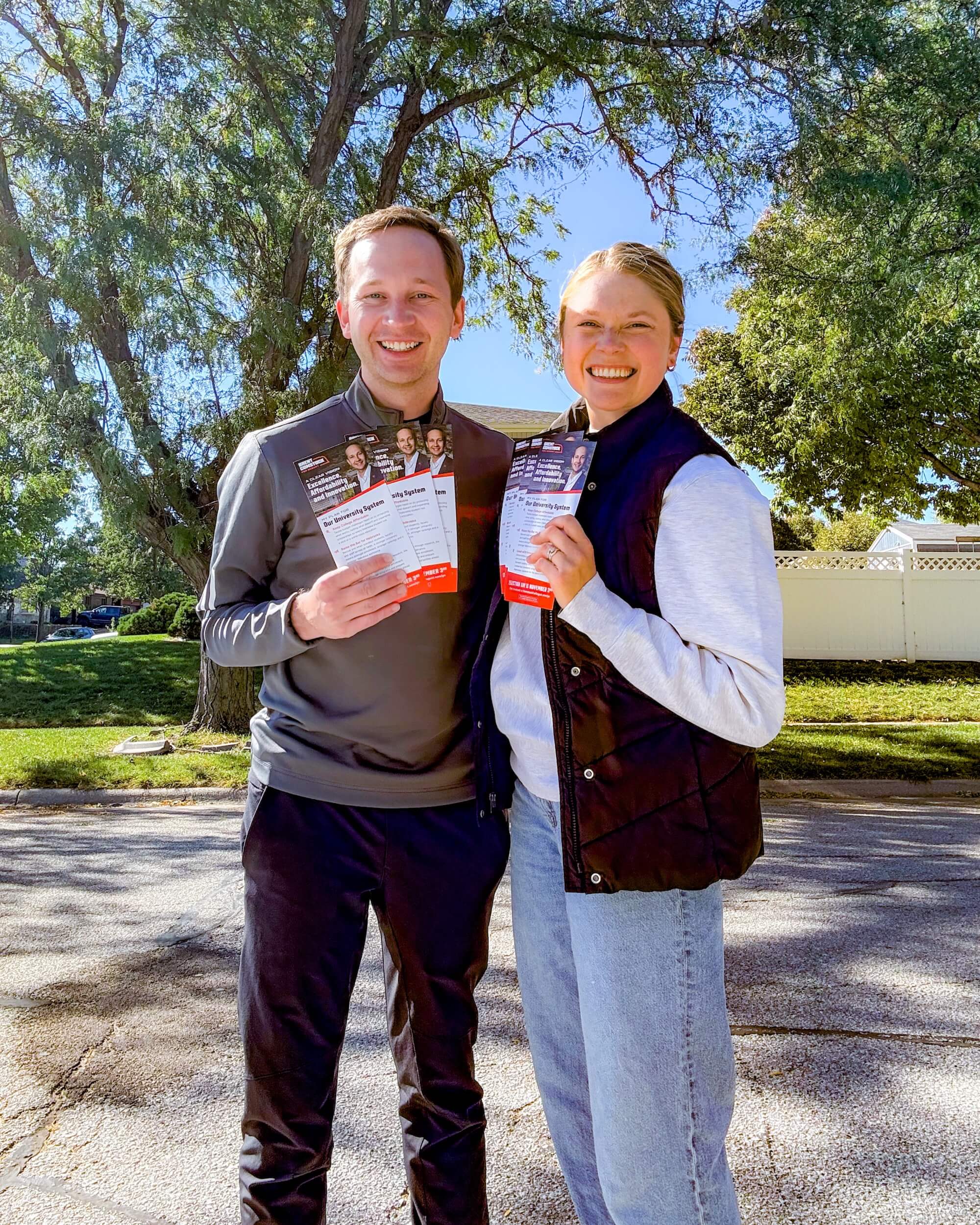 Brent Comstock and a supporter smiling outdoors on a residential street, each holding campaign flyers